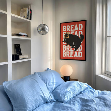 Funny wall art print of a fluffy black cat with a slice of bread on its head, set against a pink, red, orange-red, or blue background with repeated “BREAD” text. Humorous unframed cat artwork.